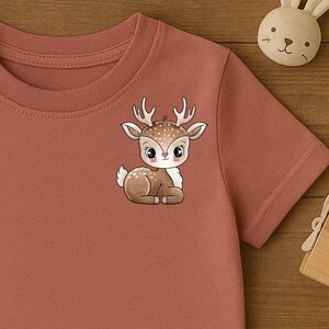 Könnte beinhalten: Ein kurzärmeliges T-Shirt mit Rundhalsausschnitt in einem gedämpften Terrakotta-Farbton. Das Shirt zeigt auf der linken Brustseite eine kleine, zentrierte Grafik eines rehähnlichen Kitzes im Cartoon-Stil mit weißen Punkten, großen Augen und Geweih.