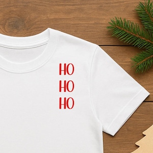 Könnte beinhalten: Weißes T-Shirt mit dem Wort "HO" dreimal in Rot. Das Bild zeigt Weihnachtsdekorationen, darunter eine rote Kugel, eine Zuckerstange, eine Geschenkbox und Tannenzweige.