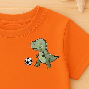 Può includere: T-shirt arancione brillante con un dinosauro dei cartoni animati che gioca a calcio. L'immagine include pantaloncini di jeans blu e scarpe da ginnastica beige. Stelle marine e stelle di legno.