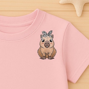 Può includere: T-shirt rosa chiaro con un disegno di capibara con una fascia fantasia. L'immagine comprende pantaloncini di jeans blu e scarpe da ginnastica beige. Sono visibili anche decorazioni in legno a forma di stella e stella marina.