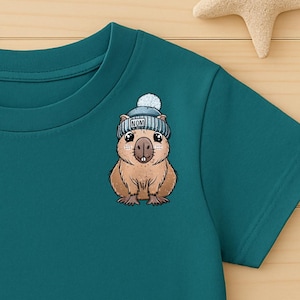 Può includere: T-shirt color verde acqua con un capibara ricamato che indossa un berretto blu e bianco. La maglietta è abbinata a pantaloncini di jeans e scarpe da ginnastica beige. Una stella e una stella marina di legno sono sulla superficie di legno.