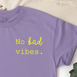 Può includere: Una maglietta viola con il testo "No bad vibes." in giallo.