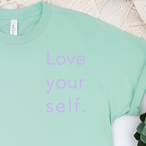Può includere: Una maglietta verde chiaro con la scritta "Love your self." stampata in viola sul davanti.