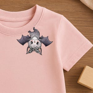 Könnte beinhalten: Hellrosa kurzärmeliges T-Shirt mit einem Fledermaus-Cartoon-Design. Die Fledermaus hat graue Flügel und eine Schleife mit Leopardenmuster. Das Shirt besteht aus weichem Material, geeignet für Kinder.