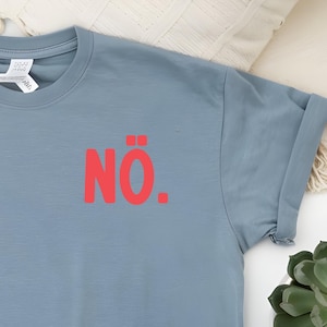 Può includere: T-shirt azzurra con maniche arrotolate. La parola "NÖ." è stampata in rosso sul petto. Questa maglietta casual è probabilmente realizzata in tessuto morbido, adatta per l'uso quotidiano.