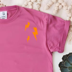 Può includere: T-shirt magenta con maniche arrotolate. Tre fulmini arancioni sono stampati sul petto sinistro. La maglietta è realizzata in materiale morbido, adatta per un abbigliamento casual. Un capo d'abbigliamento vivace.