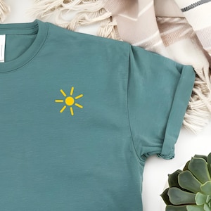 Può includere: T-shirt color verde acqua con una piccola grafica di sole giallo sulla parte superiore sinistra del petto. La maglietta ha maniche arrotolate e girocollo. La maglietta è realizzata in un materiale morbido ed è adatta per l'abbigliamento casual.