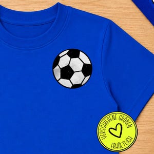 Könnte beinhalten: Ein leuchtend blaues T-Shirt mit einer schwarz-weißen Fußballgrafik auf der Brust. Ein runder gelber Aufkleber ist vorhanden, der ein schwarzes Herzsymbol und den Text VERSCHIEDENE GRÖBEN ERHÄLTLICH in schwarzer Schrift am Rand zeigt.