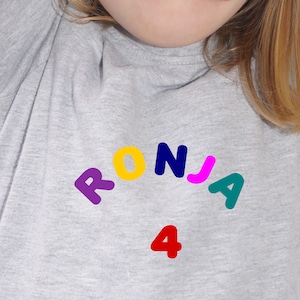 Könnte beinhalten: Ein graues T-Shirt mit dem Namen "RONJA" in bunten Buchstaben und der Zahl "4" in Rot darunter.