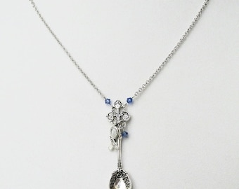 May include: A silver necklace featuring a spoon charm with a floral design and blue crystal accents. A small ribbon charm hangs from the spoon.