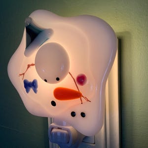 May include: A white ceramic night light shaped like a snowman with a blue hat, orange carrot nose, and black eyes. The night light is plugged into an electrical outlet.