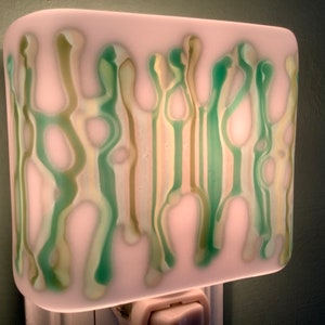 May include: A white night light with a green and brown abstract design. The light is plugged into an outlet.