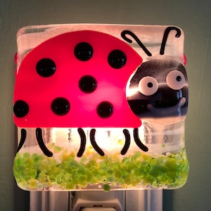 May include: A red ladybug night light with black spots and a white face. The ladybug is on a green grass background.