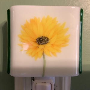 May include: A white night light with a large yellow sunflower design. The flower has a dark center and is set against a white background. The edges of the night light are trimmed with green glass.