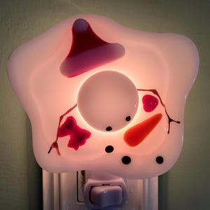 May include: A snowman-shaped night light with a red hat, carrot nose, and black eyes and mouth. The night light is white and glows with a soft, warm light. The night light plugs into a standard wall outlet.