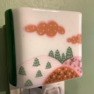 May include: A decorative night light with a landscape design. The night light features a white base with a green side and a colorful illustration of orange clouds, green trees, and pink and brown hills.