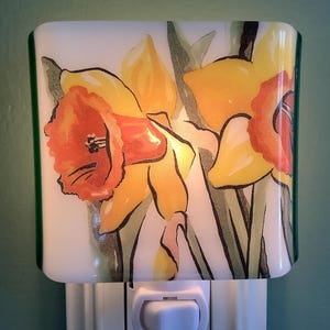 May include: A decorative night light featuring a watercolor painting of yellow and orange daffodils. The floral design is set against a white background with a green border. The night light is plugged into a white electrical outlet.
