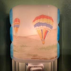 May include: A decorative night light featuring a colorful hot air balloon design. The night light has a white base with a blue trim and a landscape scene with two hot air balloons. The balloons have red, yellow, and blue stripes.