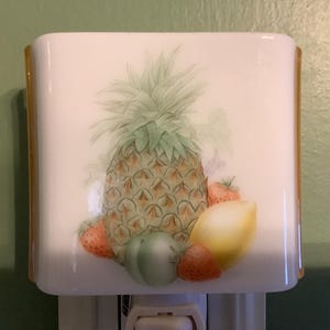 May include: A white night light with a painted design of a pineapple, strawberries, a lemon, and a green fruit. The night light has a rectangular shape with gold-colored edges. The design features a detailed illustration of the fruits.