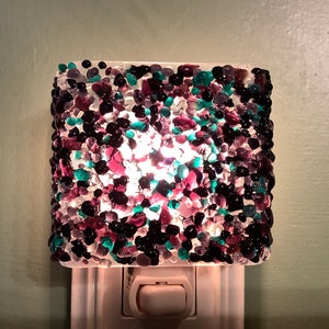 May include: A white night light with a glass shade made of small, colorful glass pieces. The shade is a mix of black, purple, and green glass pieces.