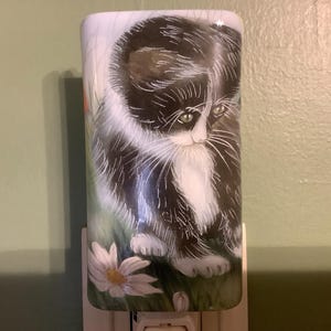 May include: A decorative night light featuring a painted image of a black and white kitten with green eyes. The kitten is depicted in a grassy area with a daisy. The night light is rectangular and has a plug-in base.
