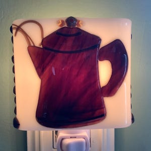 May include: A white glass nightlight with a brown and purple glass coffee pot design. The nightlight is plugged into an electrical outlet.