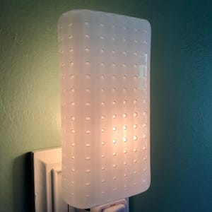 May include: A white night light with a rectangular shape and a pattern of small, evenly spaced holes. The night light is plugged into a white electrical outlet on a green wall. The light is on, emitting a soft glow.
