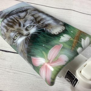 May include: Night light with a painted image of a tabby cat lying in grass with pink and white flowers. The night light has a white plastic base with an on/off switch.