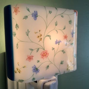 May include: A decorative night light with a floral design. The night light has a white background with a pattern of pink, blue, and yellow flowers and green leaves. The edges are trimmed with a dark blue color.