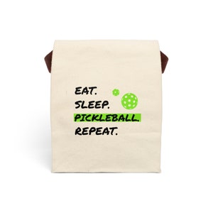May include: A white canvas tote bag with brown handles. The bag has black text that reads "Eat. Sleep. Pickleball. Repeat." and a green pickleball graphic.