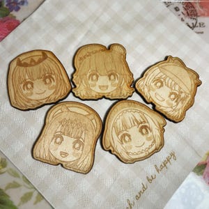 May include: Five wooden cutouts featuring smiling anime-style faces with various hairstyles. The cutouts are light brown with etched details, and are arranged on a checkered napkin. The phrase "and be happy" is visible on the napkin.
