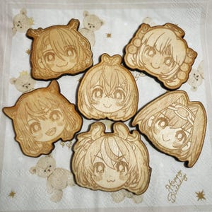 May include: Six wooden cutouts featuring anime-style character faces. Each cutout is a light brown color with detailed engraved facial features and hairstyles. The cutouts are arranged on a white napkin with a teddy bear pattern.