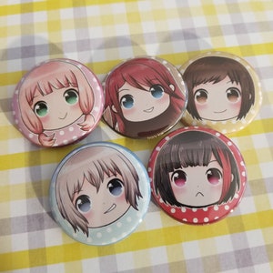 May include: Five round anime-style buttons with smiling faces. Each button features a different character with unique hair color and eye color. The backgrounds are various colors with polka dots. The buttons are on a yellow and white checkered surface.