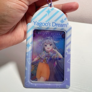 May include: A rectangular, light purple keychain with the text "Yagoo's Dream" at the top. The keychain features a colorful illustration of a smiling anime character with long hair, wearing a floral shirt and orange shorts.