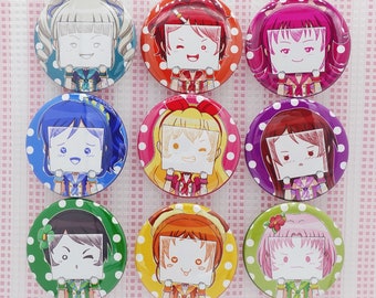 May include: Nine colorful round buttons with anime-style character illustrations. Each button features a different character with unique hair and outfit designs. The buttons have a glossy finish and are set against a pink and white striped background.