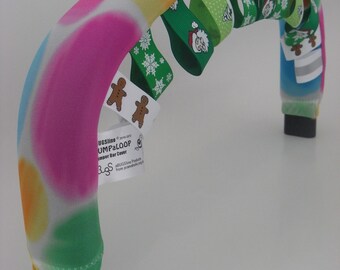 May include: A colorful, tie-dye patterned handlebar cover with a green, white, and red ribbon design. The cover is made for a stroller or a baby swing. The cover has a gingerbread man and snowflake designs on the ribbon. The text on the tag reads "BUGSline - pure fun!  JUMPALOOP Stroller Bar Cover".