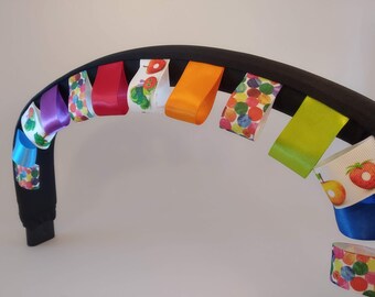Könnte beinhalten: Ein schwarzer Haarreif, der mit einer Reihe bunter Bandschlaufen verziert ist. Die Bänder zeigen einfarbige Töne wie Violett, Rot, Orange, Limettengrün und Blau sowie gemusterte Bänder mit Illustrationen von Raupen, Früchten und bunten Punkten.