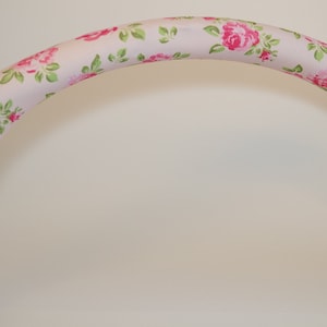 Può includere: Una copertura per il manubrio del passeggino di colore rosa e verde con motivo floreale. La copertura è realizzata in tessuto e presenta un motivo floreale con rose rosa e foglie verdi.