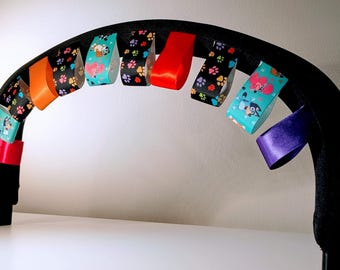 Könnte beinhalten: Eine schwarze Kinderwagenstange mit bunten Bändern daran befestigt. Die Bänder haben verschiedene Muster, darunter Pfotenabdrücke, Comicfiguren und einfarbige Farben. Die Bänder sind um die Stange gebunden und hängen herunter.