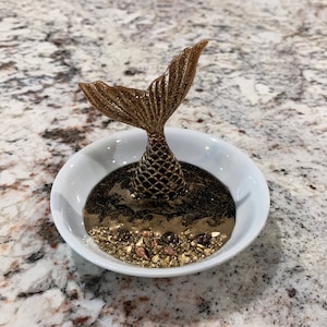 Puede incluir: Un porta anillos de cola de sirena dorada con una base de cerámica blanca y purpurina dorada. La cola está detallada con escamas y tiene un anillo descansando en la parte superior.