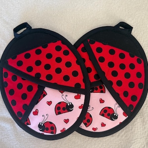 May include: Two ladybug-themed pot holders. Each pot holder has a black handle and a red and black polka-dot pattern. The bottom section features a pink background with ladybugs and hearts.