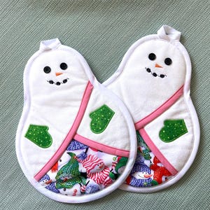 May include: Two white snowman-shaped pot holders with black button eyes, embroidered smiles, and orange carrot noses. Each has a green mitten applique and a pink accent. The bottom section features a snowman print.