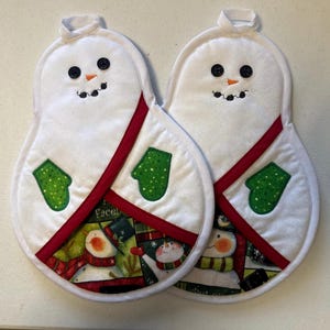 May include: Two white fabric oven mitts shaped like snowmen, featuring black button eyes, orange stitched noses, and green mittens. The lower sections display a colorful printed pattern of snowmen, and each mitt has a red fabric trim.