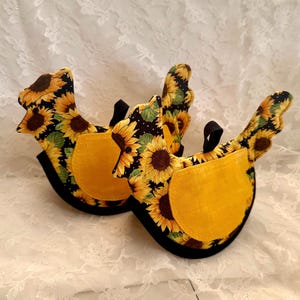 May include: Two chicken-shaped pot holders. The pot holders are decorated with a sunflower pattern on a black background. Each has a yellow pocket and a black handle. The pot holders are designed to protect hands from heat.