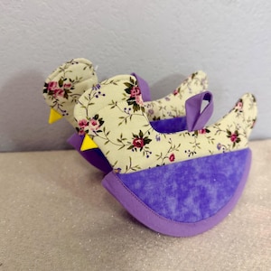 May include: Two bird-shaped fabric pot holders. The top portion features a cream-colored floral pattern with pink and burgundy roses. The bottom is a textured purple fabric. The birds have purple wings and yellow beaks. The pot holders are designed to protect hands from heat.