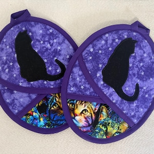 May include: Two purple and white cat-themed pot holders. Each has a black cat silhouette on a purple starry background. The bottom section features a colorful cat face design. Each pot holder has a purple loop for hanging.