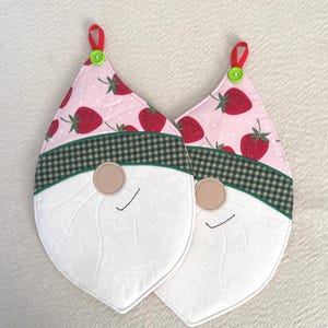 May include: Two pink and green pot holders shaped like gnomes with strawberry print and white faces. The gnomes have green and white checkered hats and a green trim around the edges.