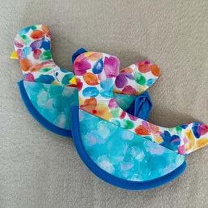 May include: Two bird-shaped pot holders. The top portion features a white background with colorful watercolor floral patterns. The bottom is a light blue with a darker blue trim. The birds have yellow beaks.