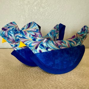 May include: Pair of blue fabric booties with a swirling pattern in shades of blue, purple, and gold. The booties have a solid blue base and a yellow triangle accent. The booties are designed to resemble birds.