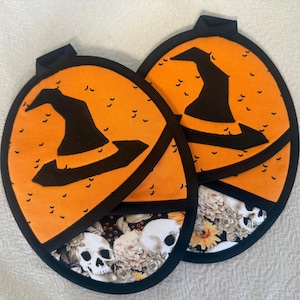 May include: Two round oven mitts featuring an orange upper section with a black witch hat graphic and small bat silhouettes, and a lower section with a floral pattern including white skulls. Each mitt has a black fabric trim and a hanging loop.
